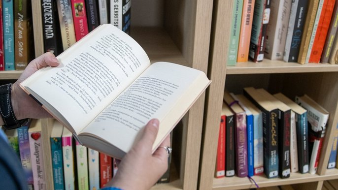 Biblioteczka przy ul. Objazdowej 17. Biblioteczka przy ul. Objazdowej 17. - Dłonie trzymające otwartą książkę na tle regału bibliotecznego.